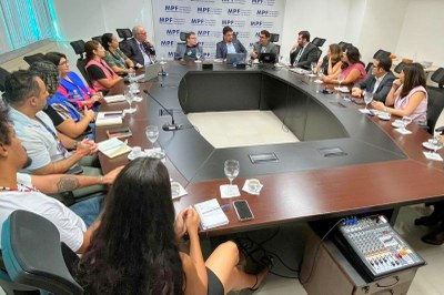 Foto: Comunicação MPF/AL Em uma sala de reuniões, cerca de 15 pessoas estão sentadas ao redor de uma grande mesa oval. Elas conversam e tomam notas, com copos de água e xícaras à frente. No fundo, há um painel com a logomarca do MPF.