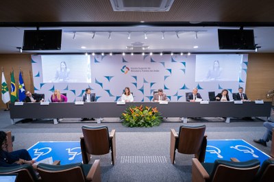Foto: Leobark Rodrigues/Secom A imagem mostra uma mesa de autoridades em um auditório, de frente para a audiência. Há sete pessoas sentadas à mesa, com um arranjo de flores no centro. Ao fundo, um painel com o logotipo da "Escola Superior do Ministério Público da União". Duas telas de TV estão posicionadas acima do painel, e uma delas exibe a imagem de uma mulher em videoconferência. Duas bandeiras estão visíveis à esquerda. No primeiro plano, há duas poltronas vazias e uma área demarcada no chão.