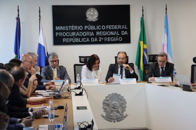 Foto de reunião formal em sala do Ministério Público Federal. Pessoas sentadas em volta de mesa, com notebooks e papéis, escutam Nicolao Dino, que fala ao microfone. No fundo, há uma placa com o nome da instituição e as bandeiras do Brasil e Estados do Rio de Janeiro e Espírito Santo.