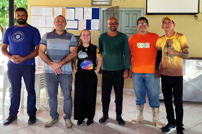 Foto: comunicação MPF Seis pessoas estão em pé, lado a lado, posando para a foto em um ambiente interno. Atrás delas há uma parede amarela com murais de avisos, porta, quadro branco, relógio e algumas fotos em moldura.