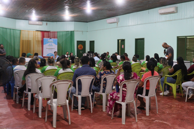 Foto mostra dezenas de pessoas, entre homens e mulheres, em ambiente de sala fechada, sentadas em cadeiras brancas de plástico, dispostas em formato de auditório; à frente, há dois homens e uma mulher sentados de frente para os demais, à frente de um banner com a inscrição "MPF na Comunidade - Escuta pública". Entre o público, há um homem de pé com um microfone na mão