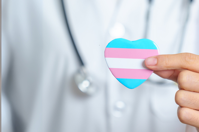 Foto mostra o torso de uma pessoa vestida com jaleco branco, com um estetoscópio no pescoço e uma mão à frente com um coração com listras azuis, rosas e branca