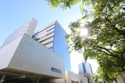 Foto: Arquivo Comunicação MPF Sede da Procuradoria Regional da República da 4ª Região vista em um ângulo de baixo para cima. A construção tem duas torres, sendo que uma delas, onde ficam os gabinetes e a maioria dos setores administrativos, a fachada é espelhada. A imagem foi registrada em um dia com céu azul, e o sol aparece por entre as folhas de uma árvore que também está enquadrada na fotografia.