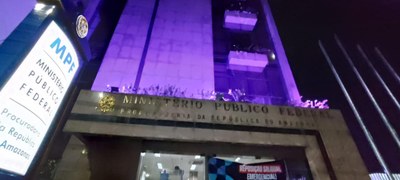 A imagem mostra a fachada de um prédio à noite, iluminado com luzes roxas. Trata-se da sede da Procuradoria da República no Amazonas, órgão do Ministério Público Federal (MPF), conforme indicado por uma placa iluminada à esquerda da imagem. A fachada do edifício é alta, com revestimento em pedra clara, e possui sacadas com plantas. Acima da entrada principal, há uma faixa com os dizeres “MINISTÉRIO PÚBLICO FEDERAL - PROCURADORIA DA REPÚBLICA NO AMAZONAS” em letras douradas. O prédio é visto de baixo para cima, acentuando sua altura. À direita da entrada, há grades metálicas verticais. Dentro do prédio, é possível ver uma área iluminada e um cartaz com os dizeres “REPOSIÇÃO SALARIAL EMERGENCIAL” em destaque.