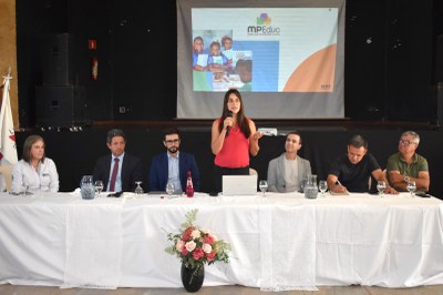 MPF-MPMG-MPEduc-realizacao-audiencia-publica-em-Piumhi-2025-08-27-Polyana-Jeha A foto mostra um grupo de seis pessoas sentadas atrás de uma mesa longa e coberta com uma toalha branca. Uma mulher, vestindo uma blusa vermelha e calça preta, está de pé no centro da mesa, falando em um microfone. Um projetor, ao fundo, exibe o logo da "MPF MP Educ". Há um buquê de flores cor-de-rosa em um vaso preto sobre a mesa. As outras pessoas na mesa, três homens e duas mulheres, estão sentadas e observando a mulher que fala.
