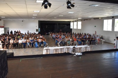 MPF-MPMG-MPEduc-realizacao-audiencia-publica-em-Piumhi-2025-08-27-plateia-auditorio Esta foto mostra uma plateia em um auditório. A plateia está lotada, com pessoas sentadas em fileiras de cadeiras voltadas para um palco de madeira. No palco, há uma mesa longa e branca com cadeiras vazias atrás. A iluminação é feita por holofotes no teto. No canto inferior direito da foto, há um homem em pé, perto do palco, usando uma vestimenta branca.