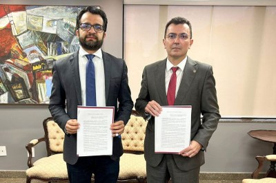 Dois homens, de terno e gravata, estão lado a lado, segurando cada um um documento assinado.