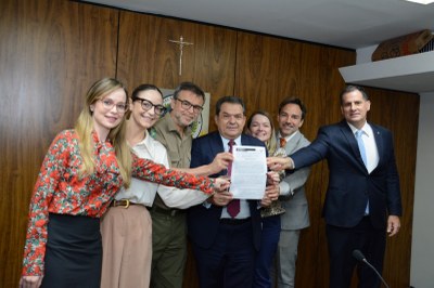 Crédito: Mauro Putini Fotografia de um grupo de sete pessoas, cinco homens e duas mulheres, em um ambiente de escritório com painel de madeira ao fundo. Eles estão de pé, lado a lado, sorrindo para a câmera. Ao centro, um homem de terno azul-marinho e gravata vinho segura um documento e os demais estendem as mãos, apoiando-o. O documento é um acordo. Todos estão vestidos com trajes formais e o clima é de celebração.