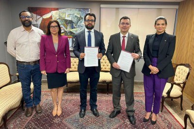 Grupo de cinco pessoas posa em uma sala, três homens e duas mulheres, todos com roupas sociais. No centro, dois homens exibem documentos.