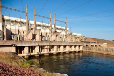 Aneel, sob licença CC BY-ND 3.0 foto de usina hidrelétrica com uma grande estrutura de concreto e turbinas em frente a um rio, com céu azul ao fundo