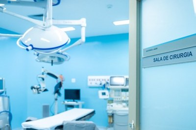A imagem mostra o interior de uma sala de cirurgia moderna e limpa, iluminada por luzes de teto azuladas. Ao fundo, está uma mesa cirúrgica vazia com lençóis brancos, cercada por equipamentos médicos de alta tecnologia, incluindo um microscópio cirúrgico, monitores e uma variedade de instrumentos. A cena é focada na porta, que tem uma placa de metal com a inscrição "SALA DE CIRURGIA", confirmando o ambiente. A foto é clara e bem iluminada, transmitindo uma sensação de profissionalismo, limpeza e esterilidade.