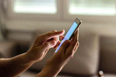 Imagem: Canva Mãos de uma pessoa seguram um smartphone na vertical, com o dedo indicador tocando a tela. A imagem mostra o uso de um celular no dia a dia.