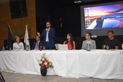 O procurador da República Gustavo Kenner Alcântara fala em audiência pública do MPEduc em Piumhi (MG), em 27/8/2025. Foto: Comunicação/MPF Esta imagem é de um grupo de pessoas sentadas atrás de uma mesa, com um dos homens em pé e falando em um microfone. Ele está usando um terno azul-marinho e gesticula com a mão direita. Os demais sentados à mesa o observam. Ao fundo, um projetor exibe uma imagem de uma ponte em uma paisagem de pôr do sol. Sobre a mesa, há um buquê de flores cor-de-rosa em um vaso preto.