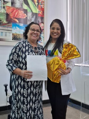 Duas mulheres, uma mais velha, a outra na faixa dos 35/30, posando em conjunto para a foto enquanto seguram um presente embrulhado em papel amarelo
