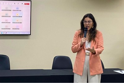Uma mulher de blazer salmão e óculos fala ao microfone em frente a uma mesa preta e uma tela que exibe participantes de uma reunião online.
