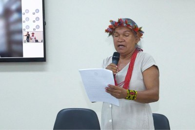 Uma mulher indígena com cocar de penas e colar vermelho lê um papel em pé, falando em um microfone, ao lado de uma tela.
