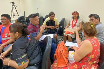 Dois grupos de pessoas, incluindo uma mulher indígena de cocar, participam de uma roda de conversa sentados em cadeiras pretas em uma sala.