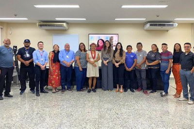 Um grupo de quinze pessoas, incluindo uma mulher indígena com cocar, posa lado a lado para uma foto em frente a uma tela, em que há rosto de outra participante do evento.