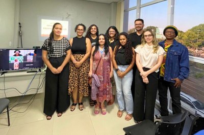  Foto de um grupo de nove pessoas, a maioria mulheres. O grupo posa sorrindo em pé em uma sala, com uma televisão e uma janela grande ao fundo. Na televisão, demais pessoas participantes do encontro.