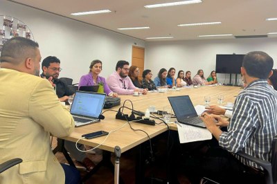 Reunião com cerca de vinte pessoas sentadas ao redor de uma mesa de conferência, algumas com notebooks abertos, em uma sala iluminada com paredes brancas e televisão ao fundo.