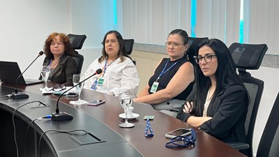 A foto mostra um enquadramento mais próximo de quatro mulheres que participam da reunião. Todas estão sentadas lado a lado à mesa, com microfones e taças d’água à frente. Elas usam crachás e trajes formais, demonstrando concentração e atenção ao que está sendo discutido. O fundo da imagem mostra as janelas com persianas e a parede branca da sala de reunião.