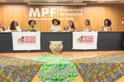 Seis mulheres negras sentadas em uma mesa de painel com faixas da "Marcha das Mulheres Negras por Reparação e Bem Viver Brasília | 2025" e um fundo com a inscrição "MPF - Procuradoria da República no Pará".