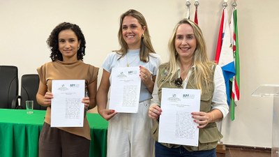 Três mulheres estão lado a lado, segurando documentos timbrados do Ministério Público Federal – Procuradoria da República em Alagoas. Elas sorriem para a câmera e ao fundo, há uma mesa com toalha verde e bandeiras do Brasil e de Alagoas, em ambiente de auditório institucional. 