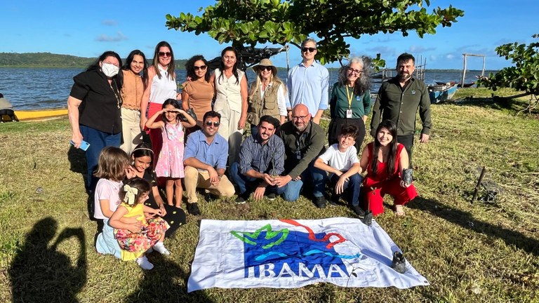 Foto em grupo, desta vez à beira de um corpo d’água, sob o sol da tarde. Há adultos e crianças, e no chão está estendida uma bandeira do Ibama. Algumas pessoas seguram mudas. A paisagem natural, com o lago e o céu azul ao fundo, reforça o caráter ambiental e colaborativo da atividade.