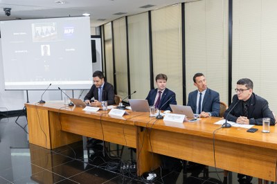 Foto de auditório onde quatro homens de terno aparecem atrás de uma bancada com microfones