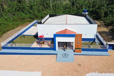 Visão aérea de uma escola nova, branca e azul, com pátio cercado, parque infantil, campo e um símbolo no chão em frente à entrada. A escola está cercada por vegetação densa.