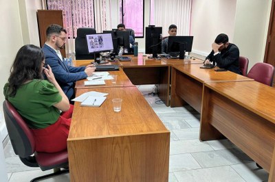 audiencia-conciliacao-processo-migrantes-2.jpg Cinco pessoas sentadas ao redor de uma grande mesa de madeira em U, em uma sala de escritório, usando computadores e papéis