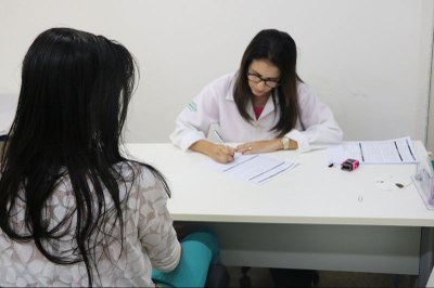 Imagem mostra a médica durante a consulta escrevendo em um papel em cima da mesa com a paciente sentada em sua frente do outro lado de uma mesa
