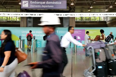 Foto ilustrativa de aeroporto com pessoas passando com malas, empurrando carrinhos, passando para o embarque.