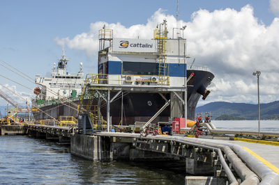 Foto: Claudio Neves/Portos do Paraná Um navio petroleiro atracado em um píer no Porto de Paranaguá. A estrutura do cais inclui um posto de controle (com o logotipo da Cattalini) e uma série de tubulações de metal para carregamento ou descarregamento de líquidos. Há montanhas e o mar ao fundo.