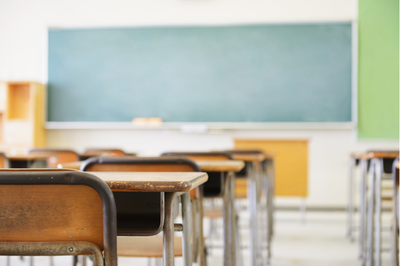 Foto ilustrativa: Canva Foto em perspectiva de uma sala de aula vazia com carteiras de madeira alinhadas e um quadro-negro desfocado ao fundo.
