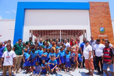 Fotos: município de São Miguel do Guamá Um grupo de crianças uniformizadas de azul, com adultos ao redor, posa na frente de um prédio escolar novo, com faixas em azul e branco. No canto direito da fachada, há um escudo e a inscrição "EMEIF Quilombola Profª. Raimunda Lopes"