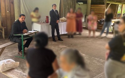Foto: MPF Foto de uma reunião em um espaço semiaberto e rústico (possivelmente uma aldeia ou galpão), com um grupo de mulheres indígenas e três homens, dois deles vestidos formalmente de terno. As mulheres estão em pé ou sentadas em bancos de madeira. A cena é focada em uma interação e diálogo entre os grupos.