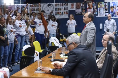 Foto: Antonio Augusto/Comunicação MPF Grupo de pessoas em auditório; mulheres de camiseta branca levantam os punhos em manifestação diante de uma mesa com autoridades sentadas e em pé. Banners e fotos de vítimas estão afixados ao fundo.