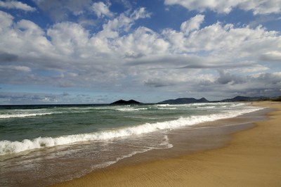 Campeche/Florianópolis/SC. Foto: Caio Vilela/MTur/Flickr MTur Destinos Foto de uma praia mostra o mar com as ondas chegando na areia e ao fundo montanhas e céu com nuvens
