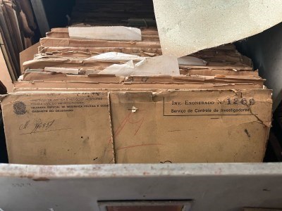 Foto: Comunicação/MPF A imagem é um close-up de uma gaveta de arquivo aberta, repleta de uma pilha alta e desorganizada de pastas de papel envelhecidas e amareladas. As pastas estão desgastadas, com bordas rasgadas e amassadas, indicando antiguidade e uso. Em uma das pastas, em primeiro plano, é possível ler parte de um carimbo e textos manuscritos com a inscrição "INV. EXONERADO Nº 1269" e "Serviço de Controle de Investigadores", sugerindo que se trata de um arquivo antigo.