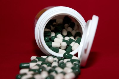 Foto ilustrativa: Canva Foto mostra um frasco de remédio aberto em cima de uma mesa e as cápsulas saindo de dentro esparramadas pela mesa vermelha