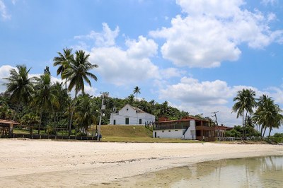Foto da Capela Nossa Senhora das neves, na Ilha de Maré, em Salvador; a igreja está situada na beira da praia com coqueiros ao redor 