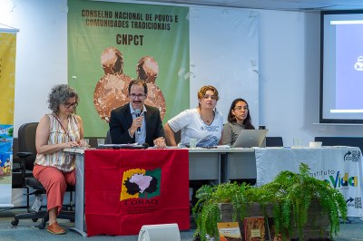Foto: Zeca Ribeiro/Comunicação MPF Quatro pessoas estão sentadas em uma mesa durante uma reunião do Conselho Nacional de Povos e Comunidades Tradicionais (CNPCT). Atrás delas há um banner verde com o nome do conselho e, à frente, bandeiras e materiais de apoio sobre a mesa.