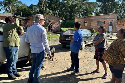Cinco pessoas reunidas em uma área externa. Ao fundo, um automóvel e duas residências de alvenaria com tijolos expostos.