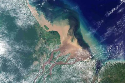 Foto: Landsat/Nasa Foto de satélite mostra a foz do rio Amazonas. A pluma marrom do rio desagua no Oceano Atlântico, azul-escuro. A terra ao redor é verde, com algumas nuvens brancas.
