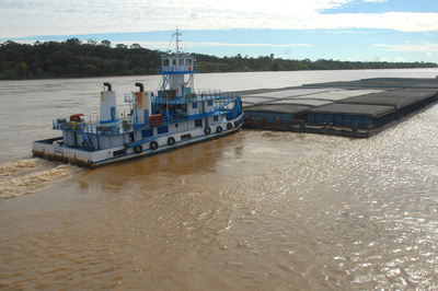 Transporte hidroviário de cargas no Rio Madeira, em Porto Velho (RO). Imagem ilustrativa. Foto: Antaq. Fotografia de um barco empurrando uma balsa com cargas em um rio com uma das margens com vegetação.
