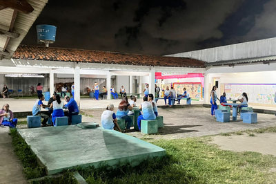 fotografia dos alunos adultos uniformizados sentados em mesas e bancos de cimento fazendo suas refeições