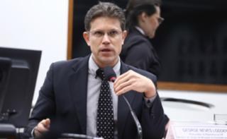 A imagem mostra uma audiência pública ou reunião de comissão em um ambiente formal. Em primeiro plano, mesas de participantes com laptops abertos (vistos de trás). Em segundo plano, quatro pessoas (três homens e uma mulher) sentadas à mesa de honra, uma delas falando ao microfone. No fundo, há uma tela exibindo um participante remoto e bandeiras ao lado de um retrato emoldurado na parede. O ambiente é de trabalho legislativo ou debate organizado.