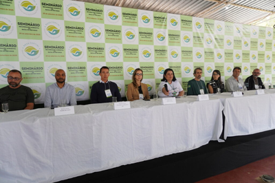 Foto: Mariana Duarte/Ascom Rosa Fortini Foto de um painel de oito participantes (seis homens e duas mulheres) está sentado em uma longa mesa coberta por toalha branca, em frente a um banner de fundo (backdrop) que repete a marca de um "Seminário de Mobilização Ambiental na Bacia do Rio Doce". Todos usam crachás.