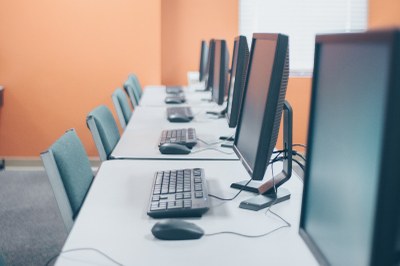 Crédito: Canva A imagem mostra uma sala de informática ou laboratório de computadores.
Há uma fileira de mesas brancas com vários monitores, teclados e mouses organizados de forma alinhada, um conjunto para cada estação de trabalho. As cadeiras são estofadas em tom azul-esverdeado e estão posicionadas de maneira uniforme diante dos computadores. O ambiente tem uma parede de cor laranja e parece bem iluminado, com luz natural entrando por uma janela com persianas no fundo.